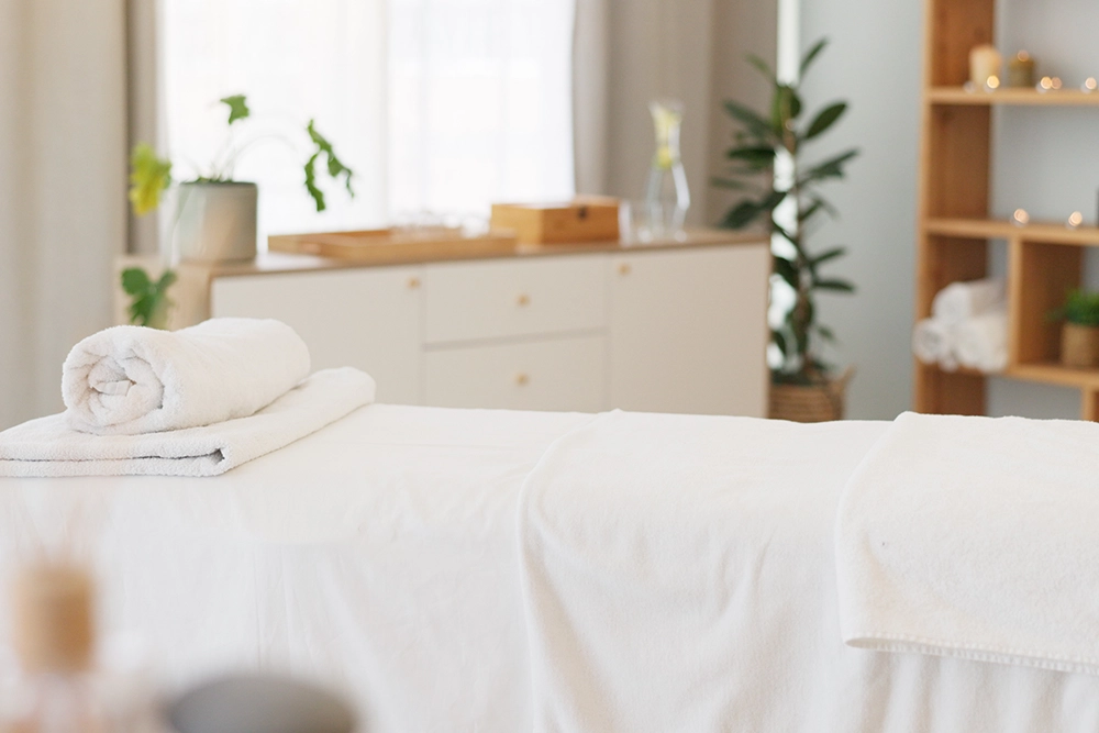 Serene massage treatment room with a white-draped table, rolled towels, potted greenery and warm natural light