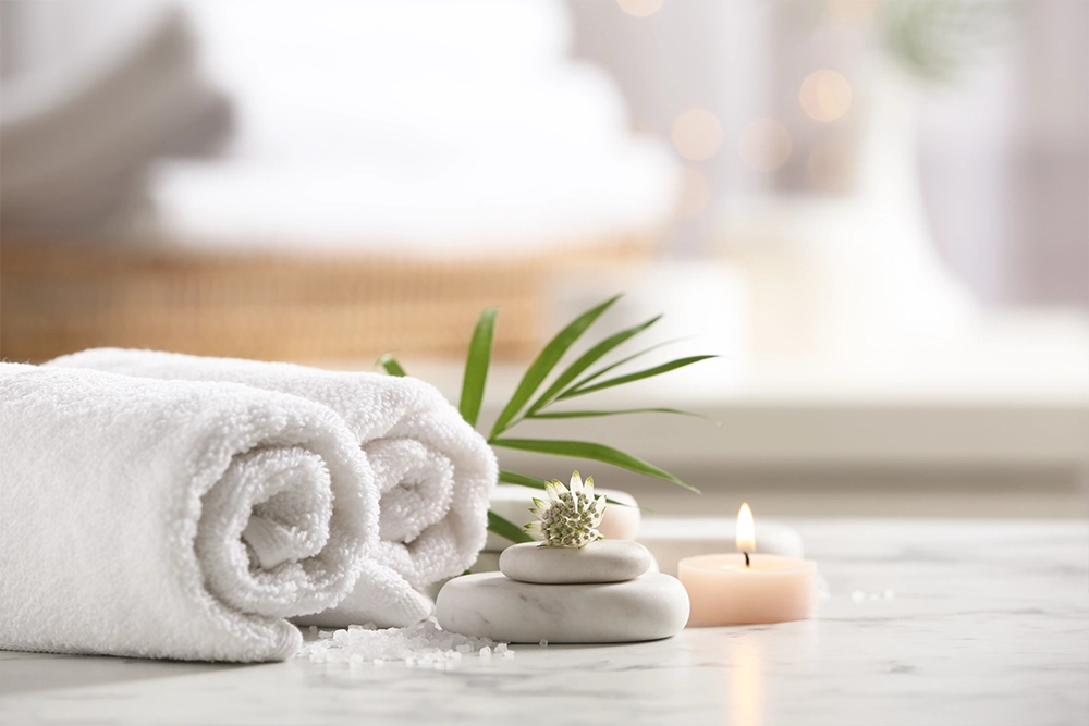 Rolled white towels, white spa stones, a lit candle and a green palm leaf on a marble surface