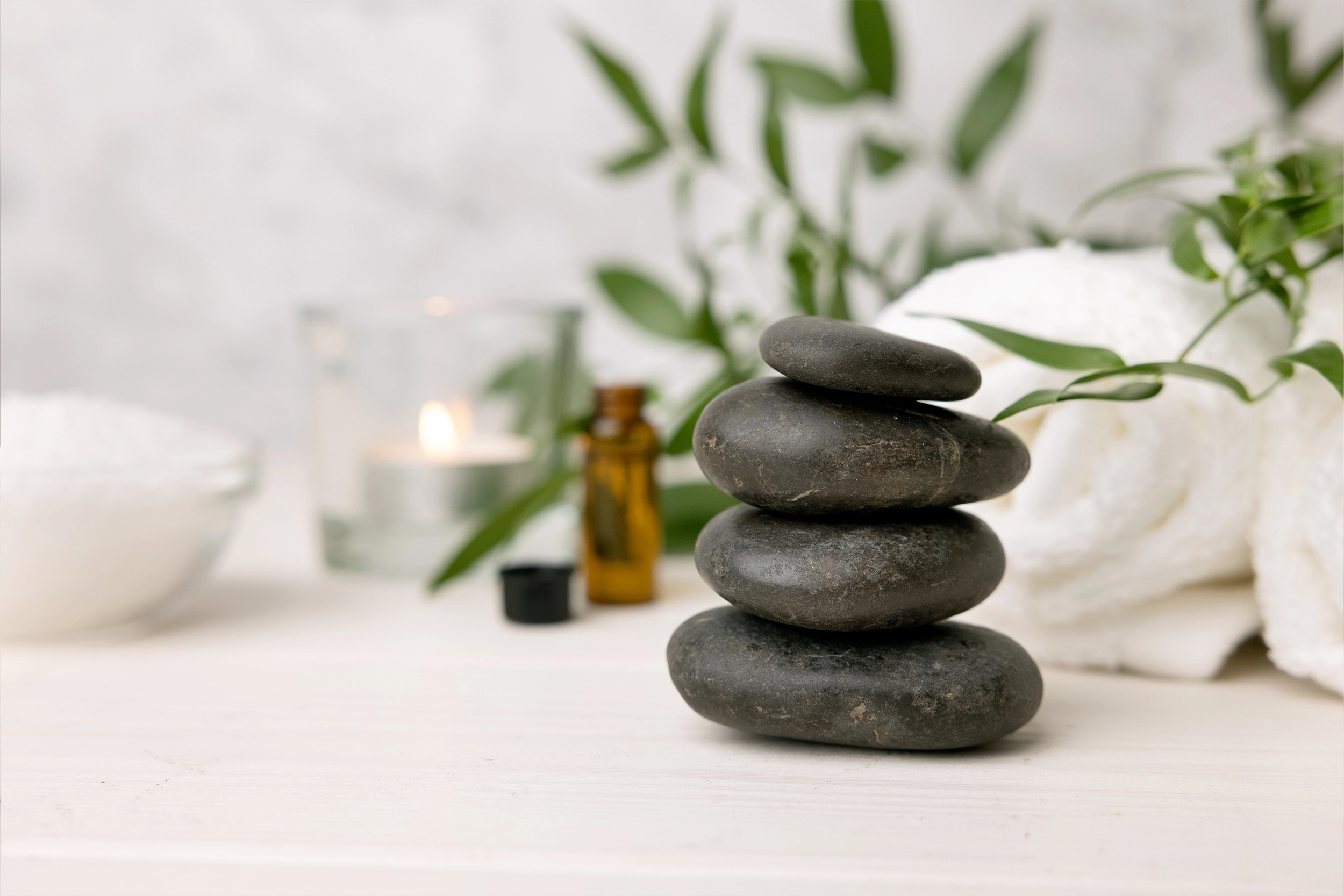 Stacked black basalt massage stones beside eucalyptus, a candle and essential oil on a white surface